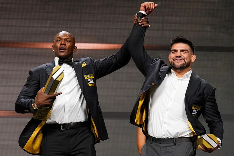 Israel Adesanya raises Kelvin Gastelum's hand at the 2025 UFC Hall of Fame induction ceremony, both in gold-lined jackets